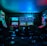 Dimly lit gaming arcade featuring a variety of brightly illuminated slot machines and video gaming screens. Neon signs and colorful displays create a vibrant atmosphere. The arrangement of gaming machines suggests an inviting space for entertainment, with chairs positioned for players.