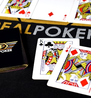 Poker-themed setup featuring a deck of cards on a table with the words 'REAL POKER' visible. Two cards, the King of Clubs and Jack of Diamonds, are prominently displayed. The design is vibrant, with detailed illustrations on the cards.