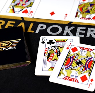 Poker-themed setup featuring a deck of cards on a table with the words 'REAL POKER' visible. Two cards, the King of Clubs and Jack of Diamonds, are prominently displayed. The design is vibrant, with detailed illustrations on the cards.