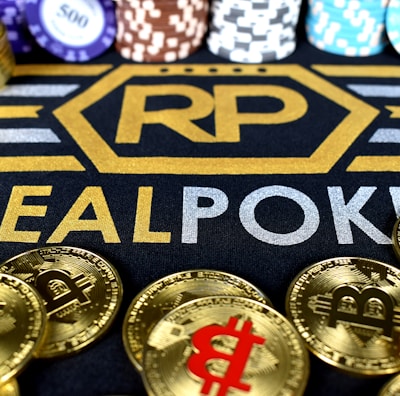 Stacks of poker chips in various colors are arranged in the background, while in the foreground, there are numerous golden coins featuring the Bitcoin symbol. The text 'REALPOKER' is prominently displayed in gold and silver lettering on a dark background.
