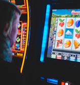 A person is closely observing a digital slot machine with colorful fruit symbols, bars, and a bell. The slot machine screen displays symbols like oranges, plums, cherries, and clouds. The background is dimly lit, creating a focused and somewhat intense atmosphere.