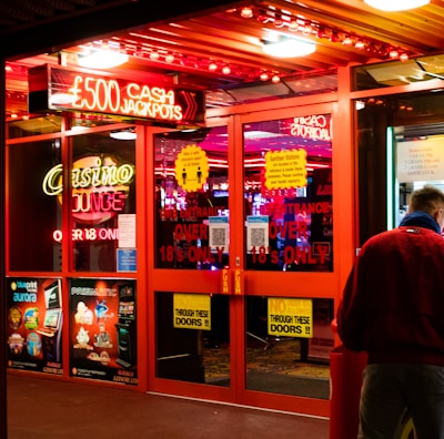 A brightly lit entrance to a casino with neon signs, including one advertising a £500 jackpot. The doors have clear signage indicating entry is restricted to those over 18. A person in a red jacket stands to the right, possibly interacting with a machine or display.