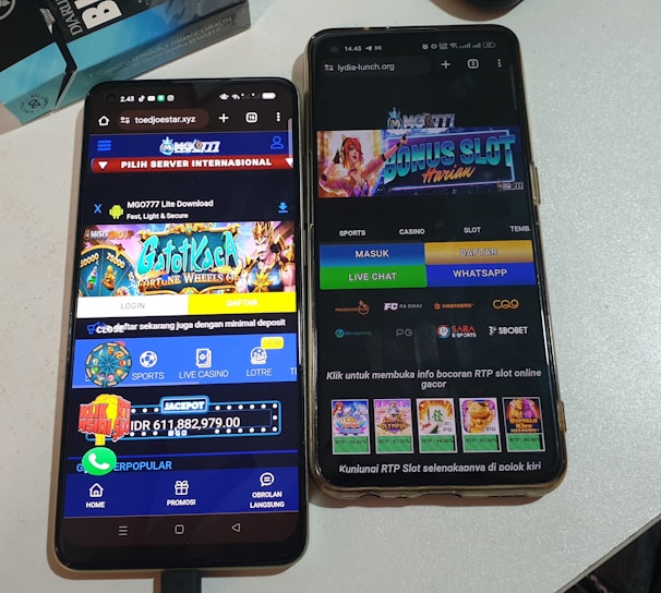 Two smartphones are displayed on a white surface, both showing gambling websites with colorful graphics, text, and various options for logging in and accessing games. The screens are bright and filled with promotional content related to casino and slot games.